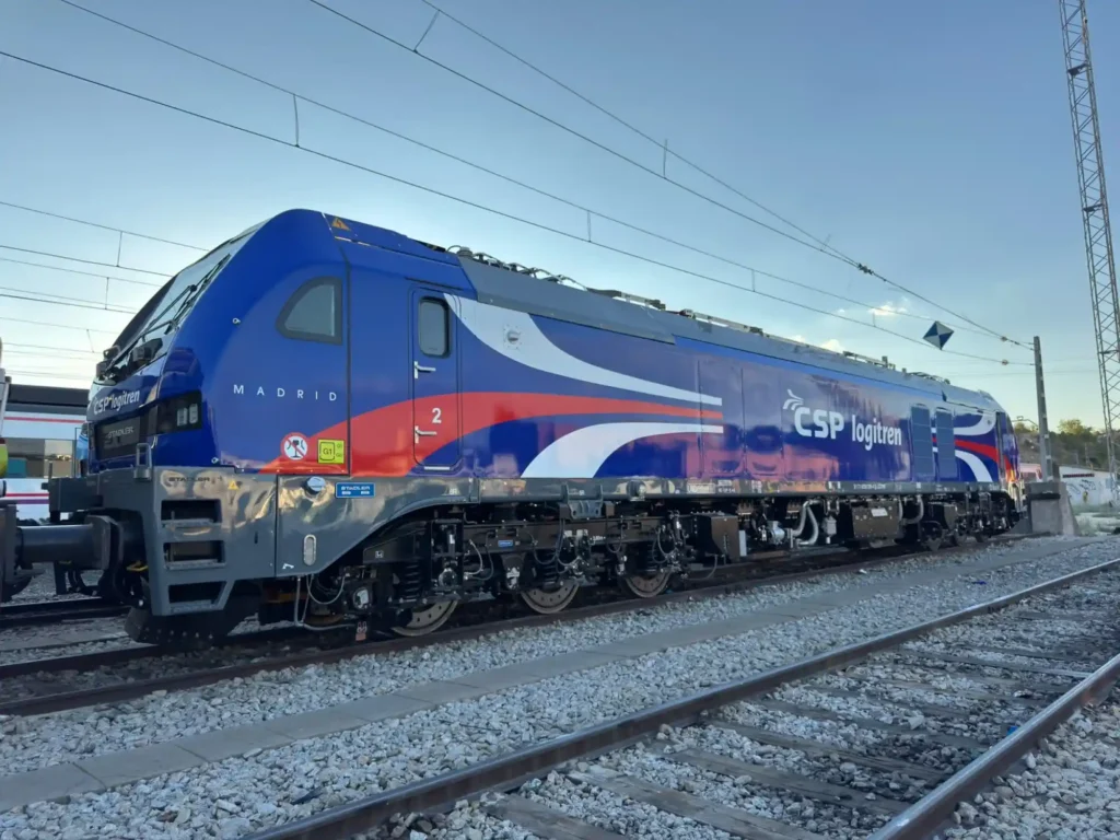 Locomotora moderna de mercancías de la compañía CSP Logitren, pintada en tonos azul, rojo y blanco, sobre vías de tren bajo un cielo despejado.