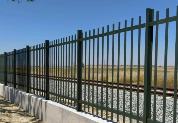 Verja metálica de barrotes verticales verdes montada sobre un muro bajo de hormigón que protege las vías del tren en un entorno rural y despejado bajo un cielo despejado.
