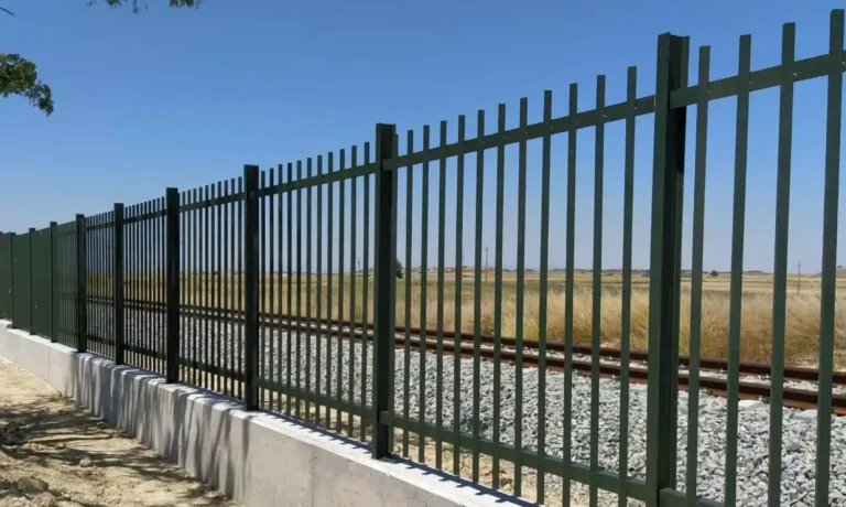 Verja metálica de barrotes verticales verdes montada sobre un muro bajo de hormigón que protege las vías del tren en un entorno rural y despejado bajo un cielo despejado.
