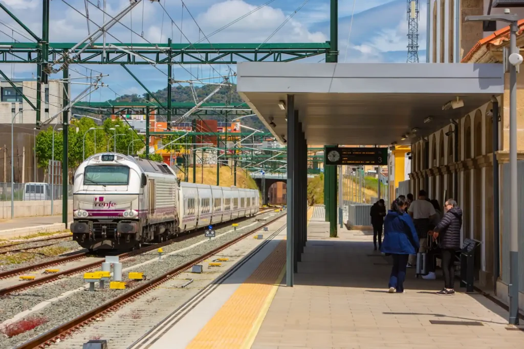 Vista del andén de la estación de trenes de Plasencia con viajeros esperando, mientras una locomotora de Renfe con vagones de pasajeros realiza su entrada por las vías bajo las estructuras metálicas de electrificación.