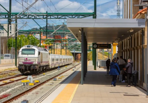 Vista del andén de la estación de trenes de Plasencia con viajeros esperando, mientras una locomotora de Renfe con vagones de pasajeros realiza su entrada por las vías bajo las estructuras metálicas de electrificación.
