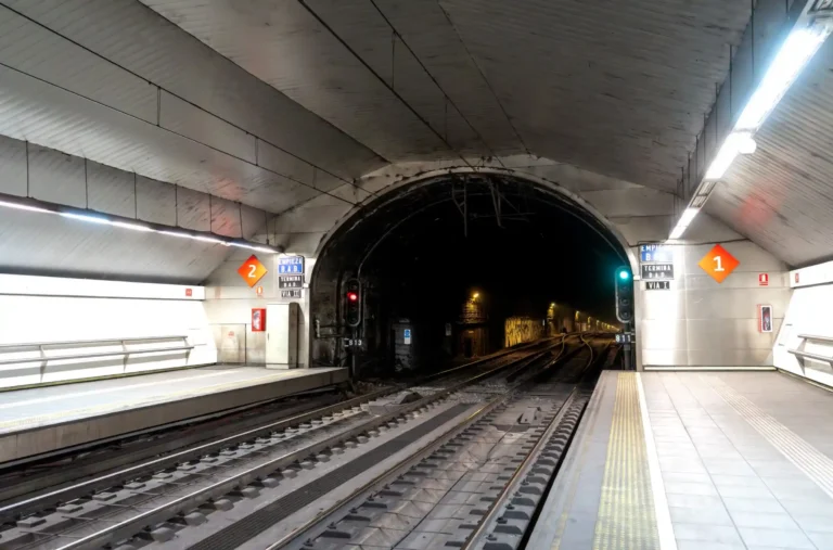Vista subterránea de los andenes y las vías del tren en la estación de Cercanías de Laguna, con la boca de un túnel al fondo y señales ferroviarias iluminadas.