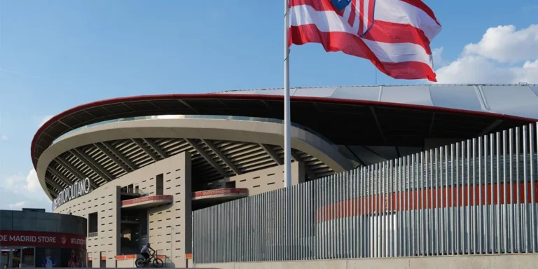 Exterior del Estadio Metropolitano en Madrid con la bandera del Atlético de Madrid y las inmediaciones de la estación de Metro Estadio Metropolitano en un día soleado.