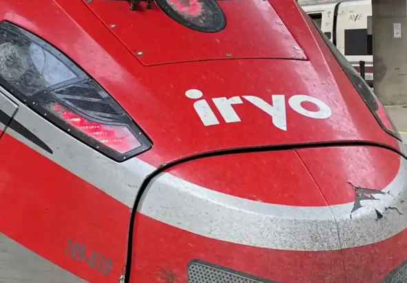 Plano detalle del morro rojo y gris de un tren de alta velocidad del operador Iryo, con el logotipo blanco visible en la parte frontal y las luces encendidas.