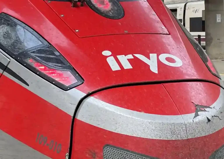Plano detalle del morro rojo y gris de un tren de alta velocidad del operador Iryo, con el logotipo blanco visible en la parte frontal y las luces encendidas.