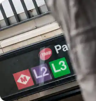 Cartel de señalización del Metro de Barcelona con el logotipo rojo de TMB y los iconos distintivos de las líneas L2 (morada) y L3 (verde), indicando el acceso a una estación de la red subterránea.