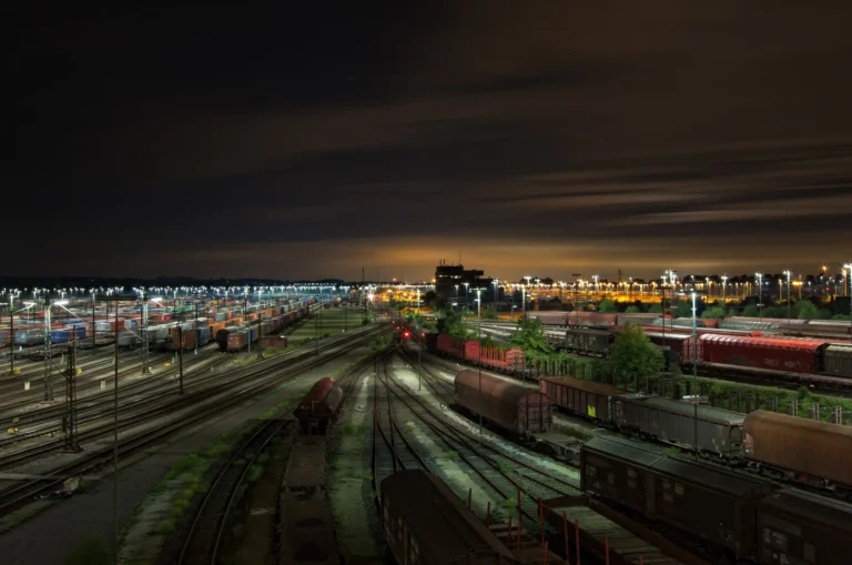 Foto nocturna aérea de una extensa y compleja playa de vías de mercancías en Europa, capturando la escala de las operaciones de carga. El cielo nocturno presenta estelas de luz, mientras que la infraestructura está iluminada por postes de luz y señalización, subrayando los desafíos de interoperabilidad que aborda la UIC Standardisation Day 2026.