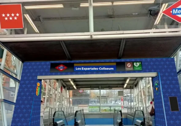 Entrada a la estación de Metro de Madrid con estructura de cristal, señalizada con el nombre 'Los Espartales-Coliseum' sobre vinilo azul con patrones y los logotipos institucionales y de Metro. Foto modificada.