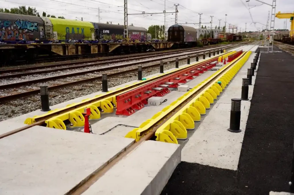 Vista en perspectiva de un cambiador de ancho automático OGI recién instalado sobre una plataforma de hormigón en la terminal de Irún, con guías laterales amarillas y un mecanismo central rojo entre los rieles, sobre un fondo de vías y vagones de mercancías.