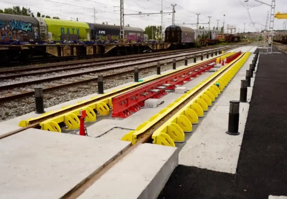 Vista en perspectiva de un cambiador de ancho automático OGI recién instalado sobre una plataforma de hormigón en la terminal de Irún, con guías laterales amarillas y un mecanismo central rojo entre los rieles, sobre un fondo de vías y vagones de mercancías.