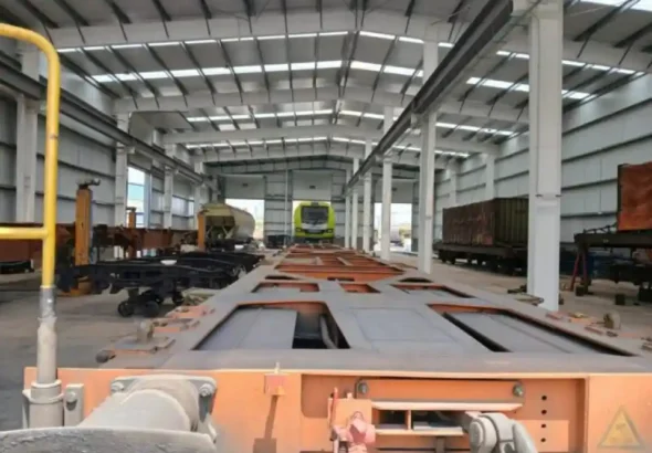 Vista interior de un amplio taller ferroviario industrial con vagones de mercancías y piezas de trenes sobre raíles.