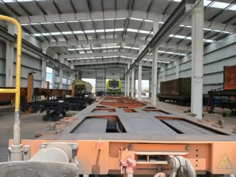 Vista interior de un amplio taller ferroviario industrial con vagones de mercancías y piezas de trenes sobre raíles.