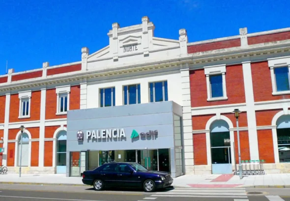 Fachada principal de la estación de tren de Palencia en un día despejado, con estructura de ladrillo rojo y detalles blancos, letreros de «Palencia» y «Adif» en la entrada y un coche oscuro aparcado enfrente.