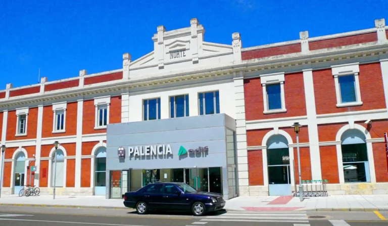 Fachada principal de la estación de tren de Palencia en un día despejado, con estructura de ladrillo rojo y detalles blancos, letreros de «Palencia» y «Adif» en la entrada y un coche oscuro aparcado enfrente.