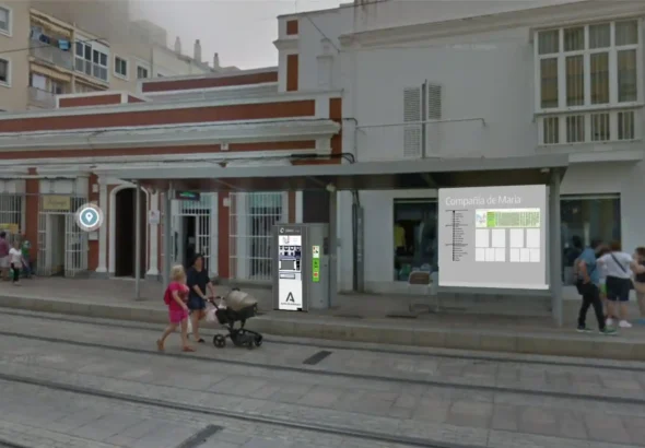 Recreación de una marquesina del Trambahía en la parada Compañía de María, provista de una máquina expendedora de billetes y un panel informativo actualizado con esquemas de línea y avisos, en una calle peatonal por la que transitan varios peatones.