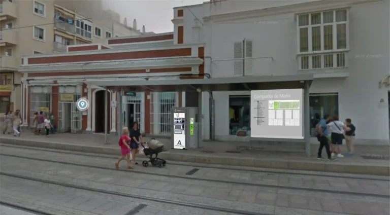Recreación de una marquesina del Trambahía en la parada Compañía de María, provista de una máquina expendedora de billetes y un panel informativo actualizado con esquemas de línea y avisos, en una calle peatonal por la que transitan varios peatones.