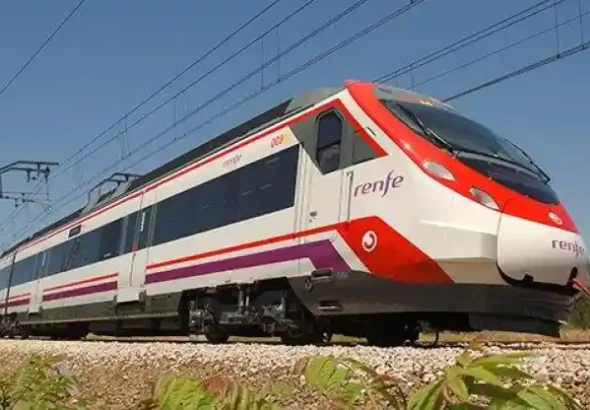 Un tren de Renfe de colores blanco, rojo y morado desplazándose por las vías sobre un balasto de piedras, bajo un cielo despejado.