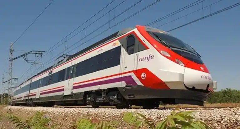 Un tren de Renfe de colores blanco, rojo y morado desplazándose por las vías sobre un balasto de piedras, bajo un cielo despejado.