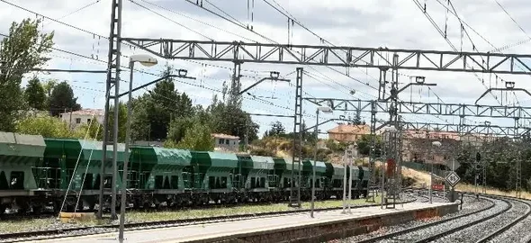 Vías de tren de la red convencional con catenarias y postes de electrificación en primer plano, junto a una hilera de vagones de mercancías verdes estacionados en una estación de ámbito rural.