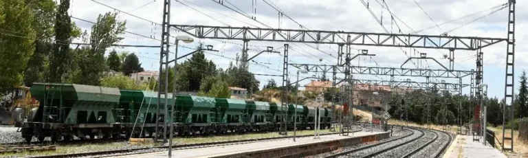 Vías de tren de la red convencional con catenarias y postes de electrificación en primer plano, junto a una hilera de vagones de mercancías verdes estacionados en una estación de ámbito rural.