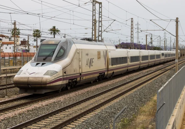 Tren de alta velocidad Renfe AVE Serie 102 de color blanco con detalles morados circulando sobre las vías a su paso por una zona con catenarias y vegetación.