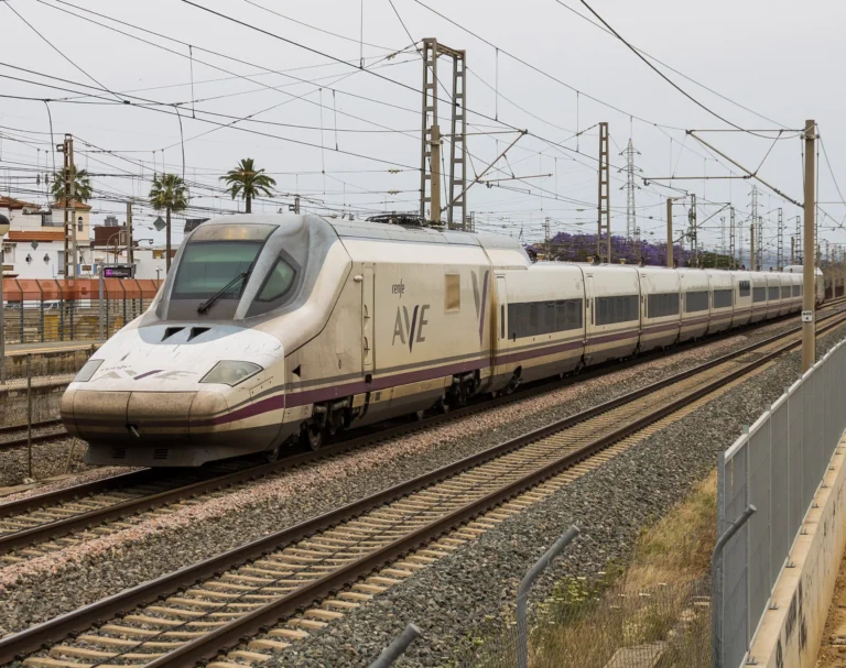 Tren de alta velocidad Renfe AVE Serie 102 de color blanco con detalles morados circulando sobre las vías a su paso por una zona con catenarias y vegetación.