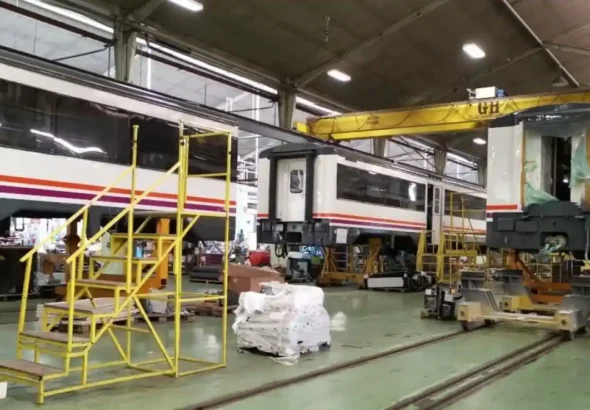 Un taller ferroviario de Renfe con vagones de tren blancos y rojos en mantenimiento, una escalera amarilla de metal en primer plano y maquinaria pesada en el fondo.