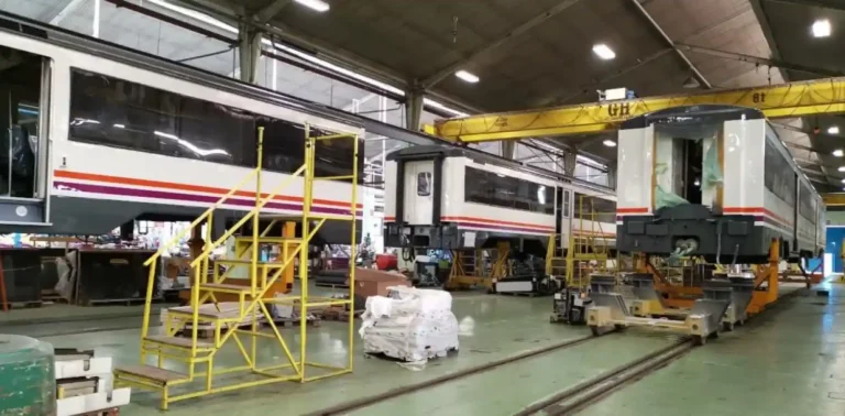 Un taller ferroviario de Renfe con vagones de tren blancos y rojos en mantenimiento, una escalera amarilla de metal en primer plano y maquinaria pesada en el fondo.
