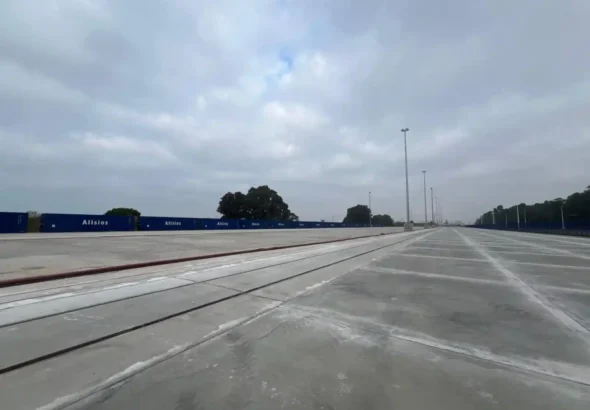 Vista en perspectiva de la explanada de hormigón pavimentada y las vías ferroviarias integradas en la terminal intermodal del Muelle Sur del Puerto de Huelva, con contenedores azules al fondo. Una de las vías pertenece al servicio de Autopista Ferroviaria.