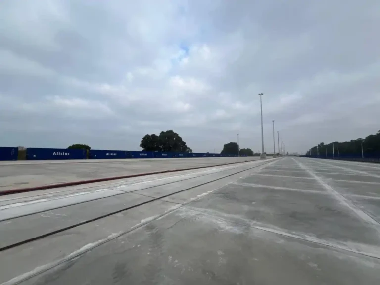 Vista en perspectiva de la explanada de hormigón pavimentada y las vías ferroviarias integradas en la terminal intermodal del Muelle Sur del Puerto de Huelva, con contenedores azules al fondo. Una de las vías pertenece al servicio de Autopista Ferroviaria.