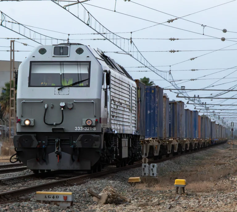 Locomotora diésel gris encabezando un largo tren de mercancías cargado con contenedores sobre la vía férrea.