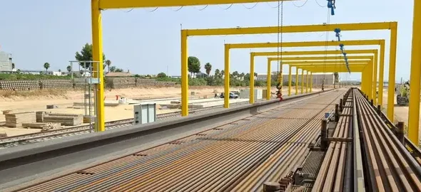 Tren carrilero cargado con largas vías de acero UIC-60 bajo pórticos amarillos en una base de montaje de vía ferroviaria de alta velocidad Murcia - Lorca.