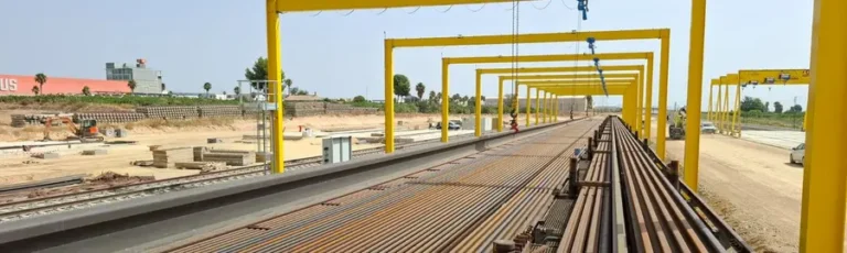 Tren carrilero cargado con largas vías de acero UIC-60 bajo pórticos amarillos en una base de montaje de vía ferroviaria de alta velocidad Murcia - Lorca.