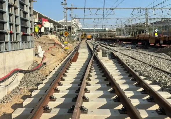 Vista en perspectiva de un desvío de vías de tren recién instaladas con traviesas de hormigón claro en la estación de Atocha, con estructuras de obra, maquinaria ferroviaria y operarios al fondo bajo la catenaria.