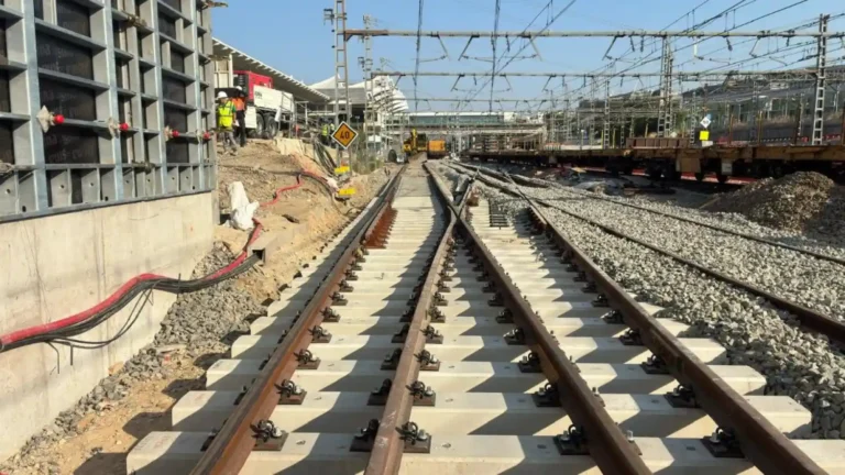 Vista en perspectiva de un desvío de vías de tren recién instaladas con traviesas de hormigón claro en la estación de Atocha, con estructuras de obra, maquinaria ferroviaria y operarios al fondo bajo la catenaria.