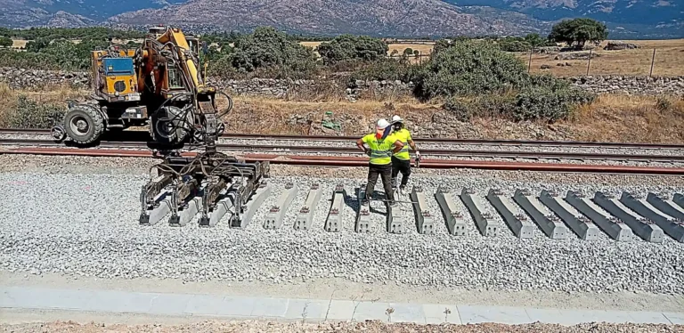 Maquinaria pesada y dos operarios instalando traviesas de hormigón sobre balasto en la plataforma de las obras del tren entre Colmenar Viejo y Soto del Real.