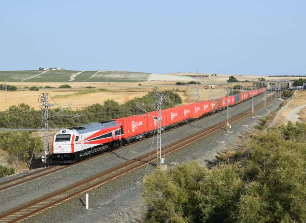 Locomotora diésel-eléctrica de gran tamaño pintada en blanco, rojo y negro con el logotipo de Boluda, arrastrando una larga hilera de vagones con contenedores marítimos rojos en una vía de doble sentido rodeada de campo.