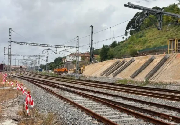 Vista de varias vías de tren en curva junto a una ladera en obras donde se observan armaduras metálicas cilíndricas inclinadas, maquinaria pesada y una pilotadora al fondo.