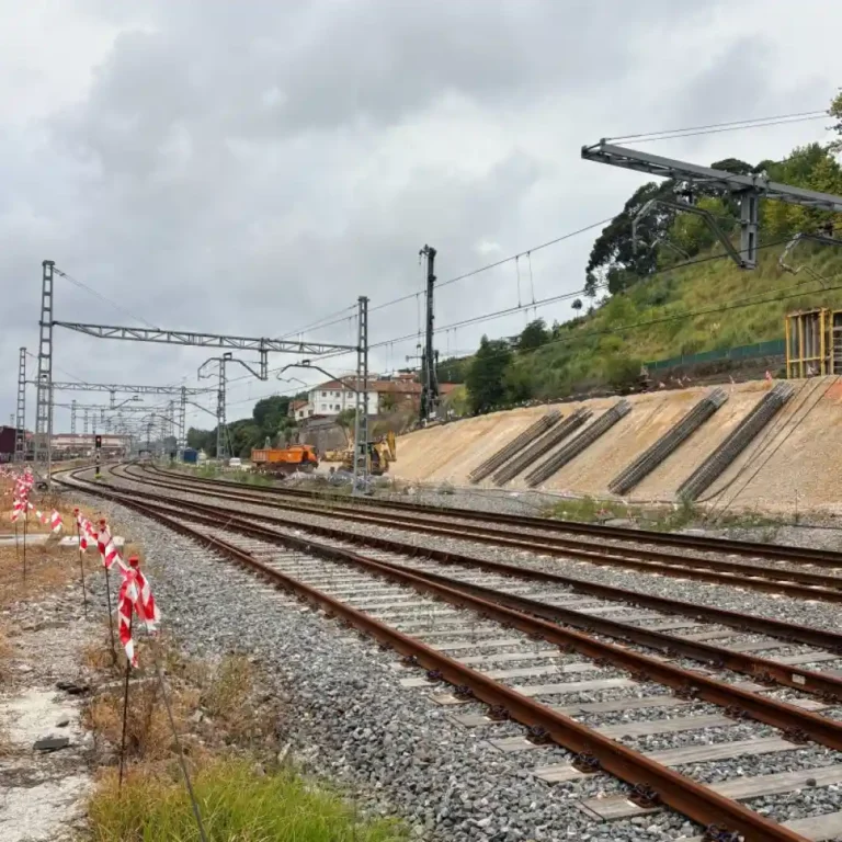 Vista de varias vías de tren en curva junto a una ladera en obras donde se observan armaduras metálicas cilíndricas inclinadas, maquinaria pesada y una pilotadora al fondo.