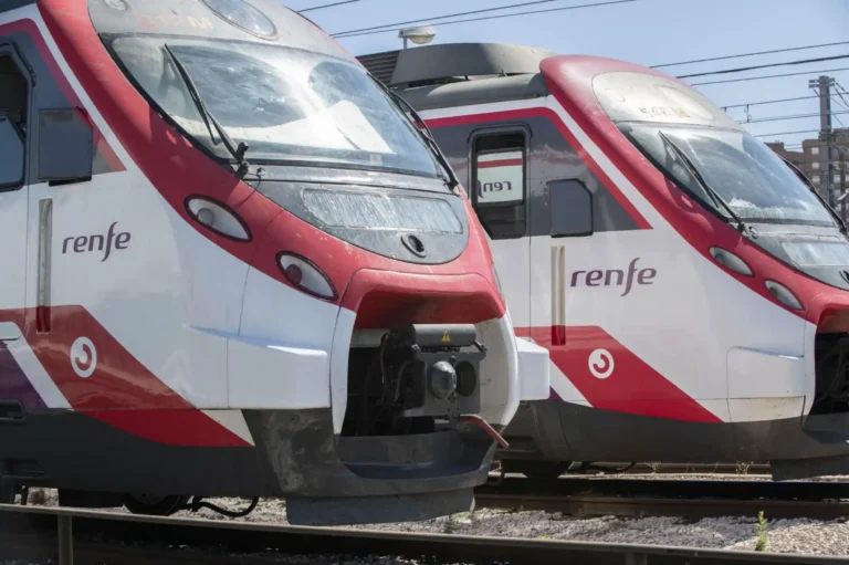 Primer plano de la parte frontal de dos trenes Civia de Cercanías Renfe de colores blanco, rojo y morado, estacionados en las vías de la estación.