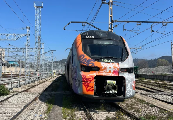 Plano frontal de un tren moderno de Cercanías Renfe con los colores naranja, blanco y gris, cubierto con pintadas y grafitis en la cabina. Se encuentra detenido en una zona de vías ferromagnticas al aire libre bajo un cielo despejado.