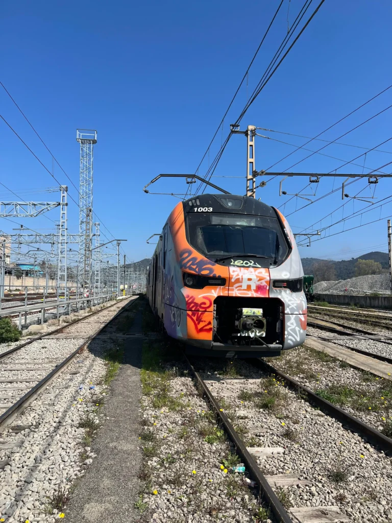 Plano frontal de un tren moderno de Cercanías Renfe con los colores naranja, blanco y gris, cubierto con pintadas y grafitis en la cabina. Se encuentra detenido en una zona de vías ferromagnticas al aire libre bajo un cielo despejado.