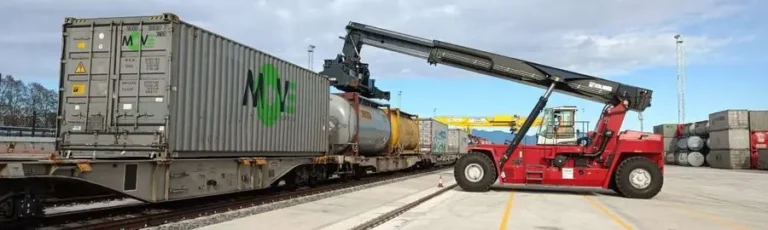 Grúa apiladora roja manipulando contenedores de transporte intermodal sobre vagones de tren en una terminal ferroviaria.