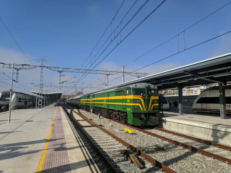 Locomotora diésel clásica de la serie 321/ALCo en tonos verde y amarillo de ALSA, estacionada en una vía de estación junto a sus coches de viajeros de los años 60.