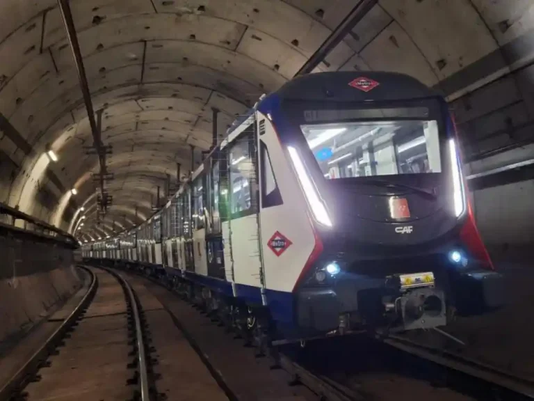 Tren moderno de Metro de Madrid de la marca CAF, con los colores blanco, azul y rojo, circulando dentro de un túnel subterráneo iluminado.