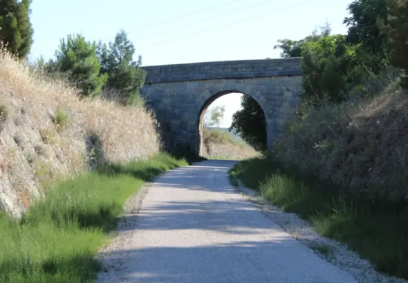 Camino rural de tierra clara flanqueado por vegetación que pasa por debajo de un antiguo puente o viaducto de arco de piedra bajo un cielo despejado.